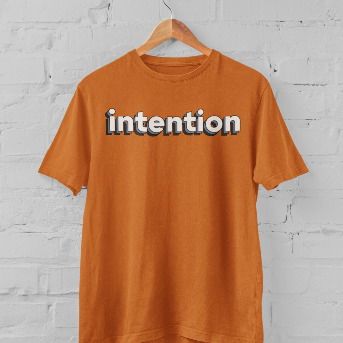a burnt orange t shirt with intention written in 3 D letters on a hanger in front of a white brick backround