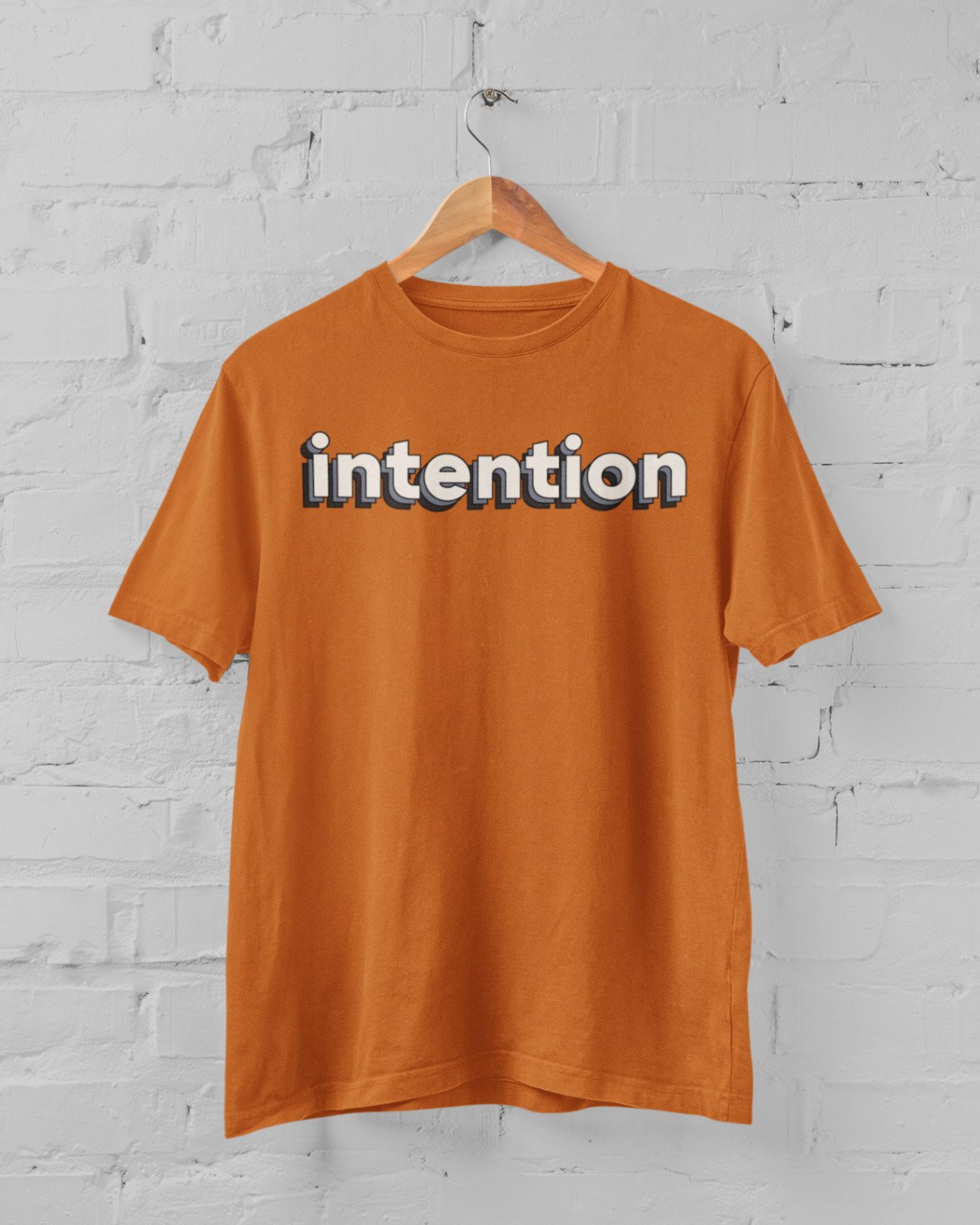 a burnt orange t shirt with intention written in 3 D letters on a hanger in front of a white brick backround