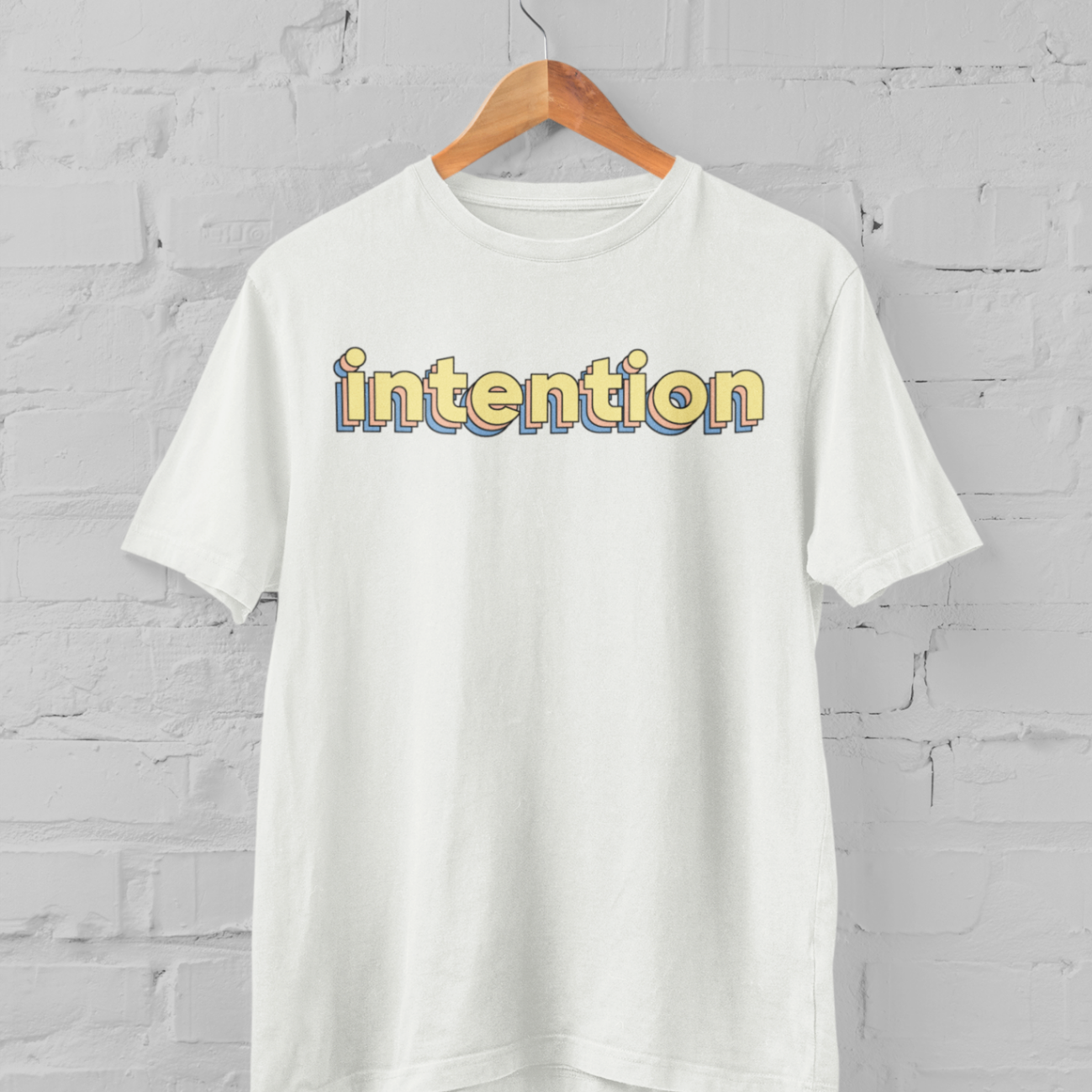 a white t shirt with intention written in 3 D letters on a hanger in front of a white brick backround