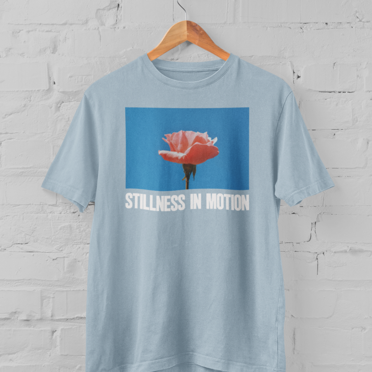 a light blue tshirt with stillness in motion written in text under a pink flower over a sky blue backround