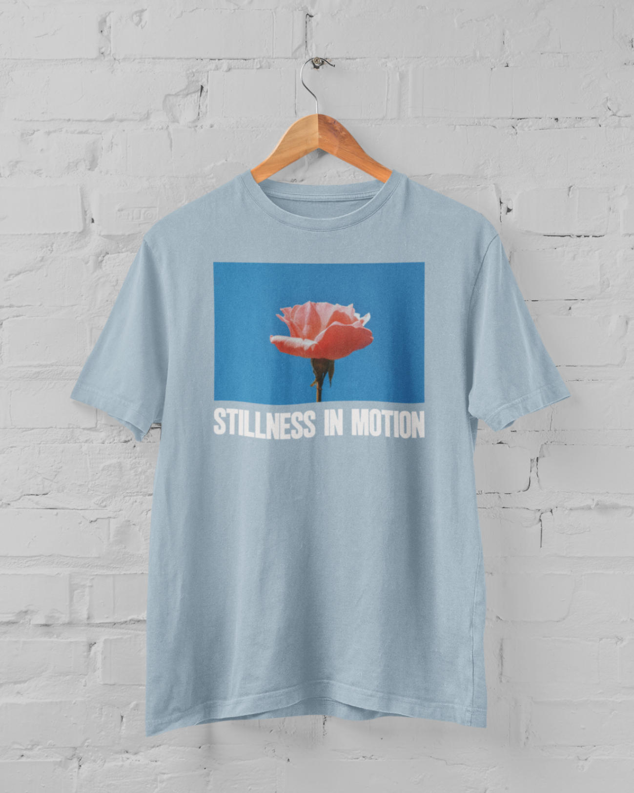 a light blue tshirt with stillness in motion written in text under a pink flower over a sky blue backround