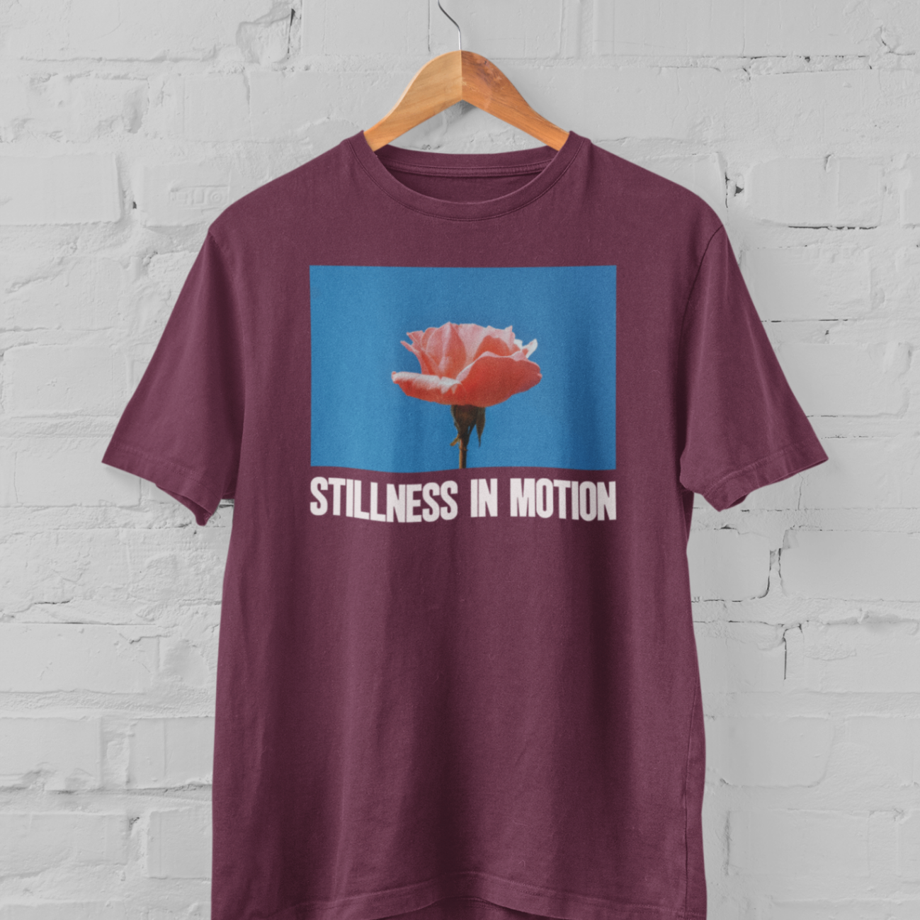 a maroon tshirt with stillness in motion written in text under a pink flower over a sky blue backround