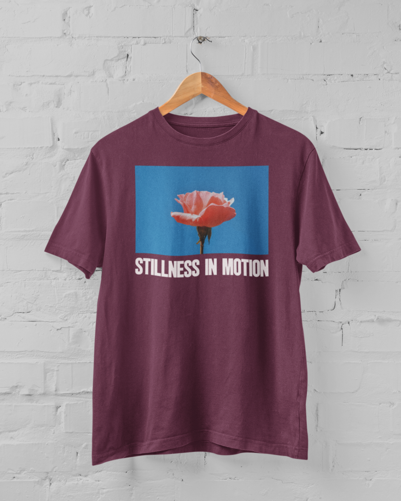 a maroon tshirt with stillness in motion written in text under a pink flower over a sky blue backround