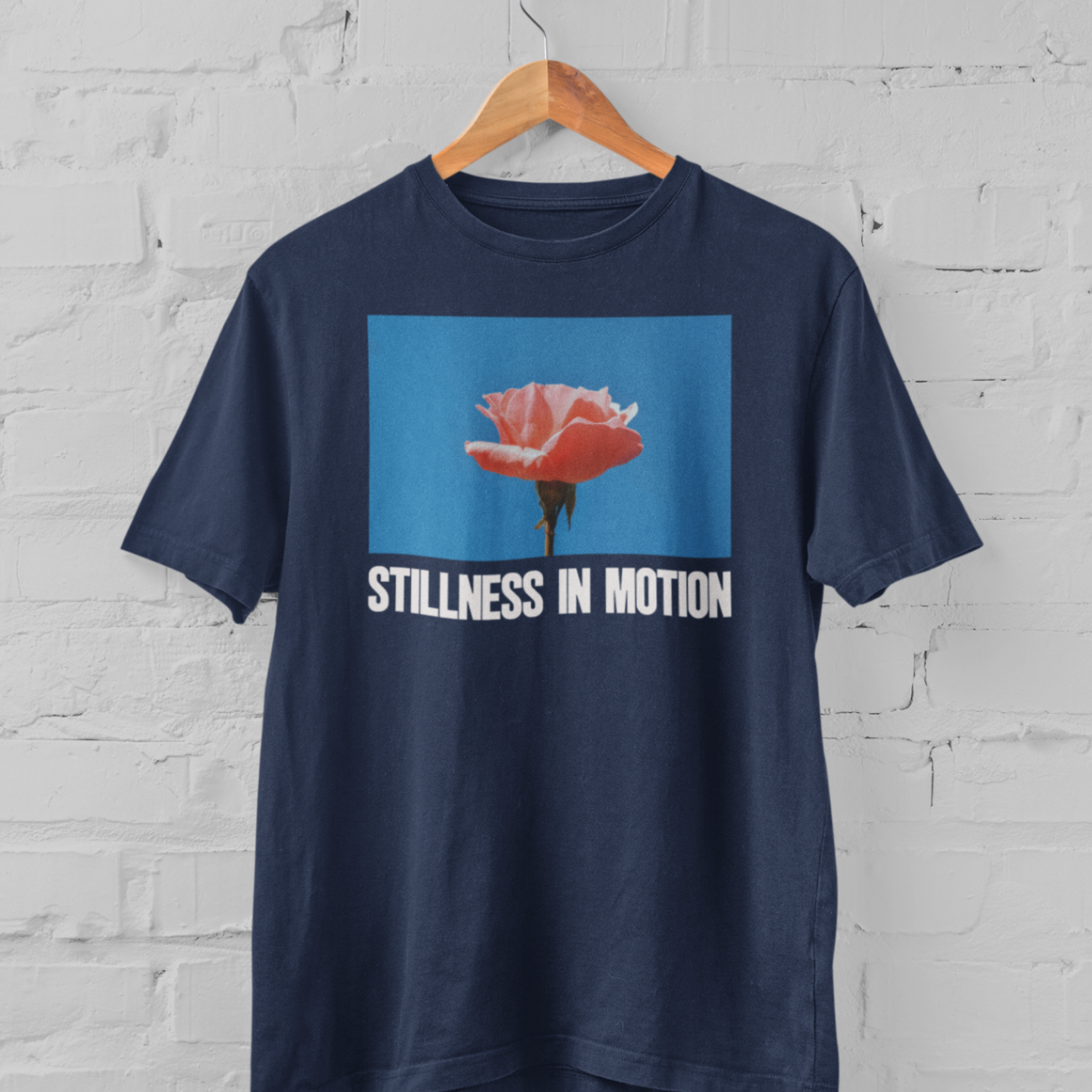 a navy tshirt with stillness in motion written in text under a pink flower over a sky blue backround