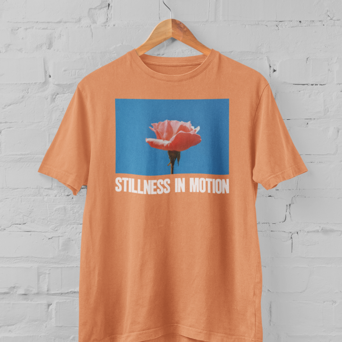 a sunset orange tshirt with stillness in motion written in text under a pink flower over a sky blue backround