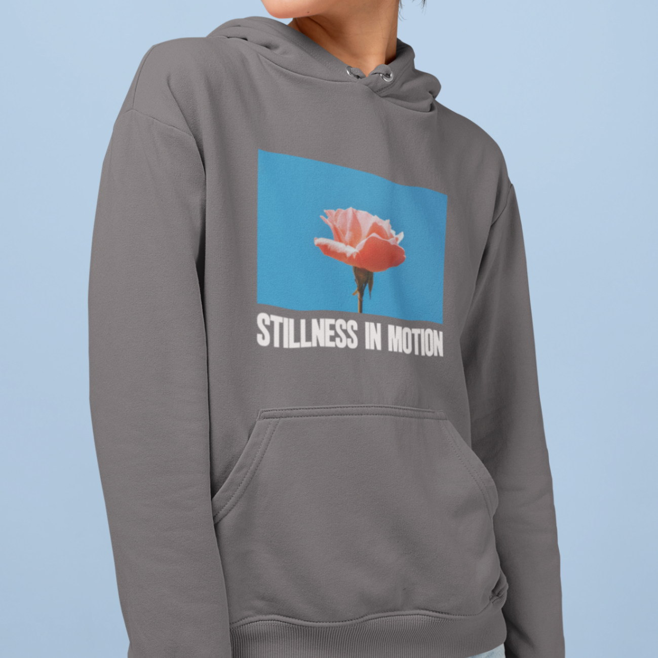 a cool woman wearing a steel grey hoodie with stillness in motion written in text under a picture of a pink flower over a sky blue backround