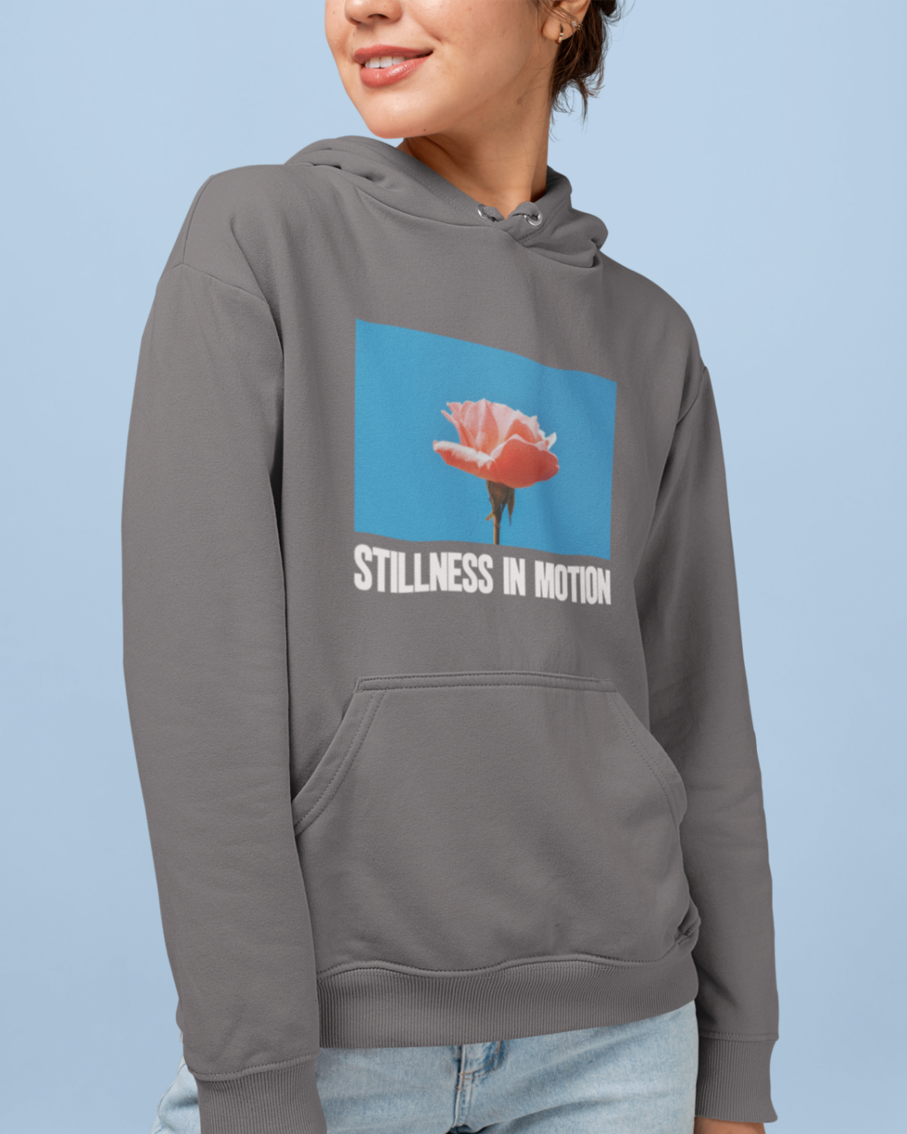 a cool woman wearing a steel grey hoodie with stillness in motion written in text under a picture of a pink flower over a sky blue backround