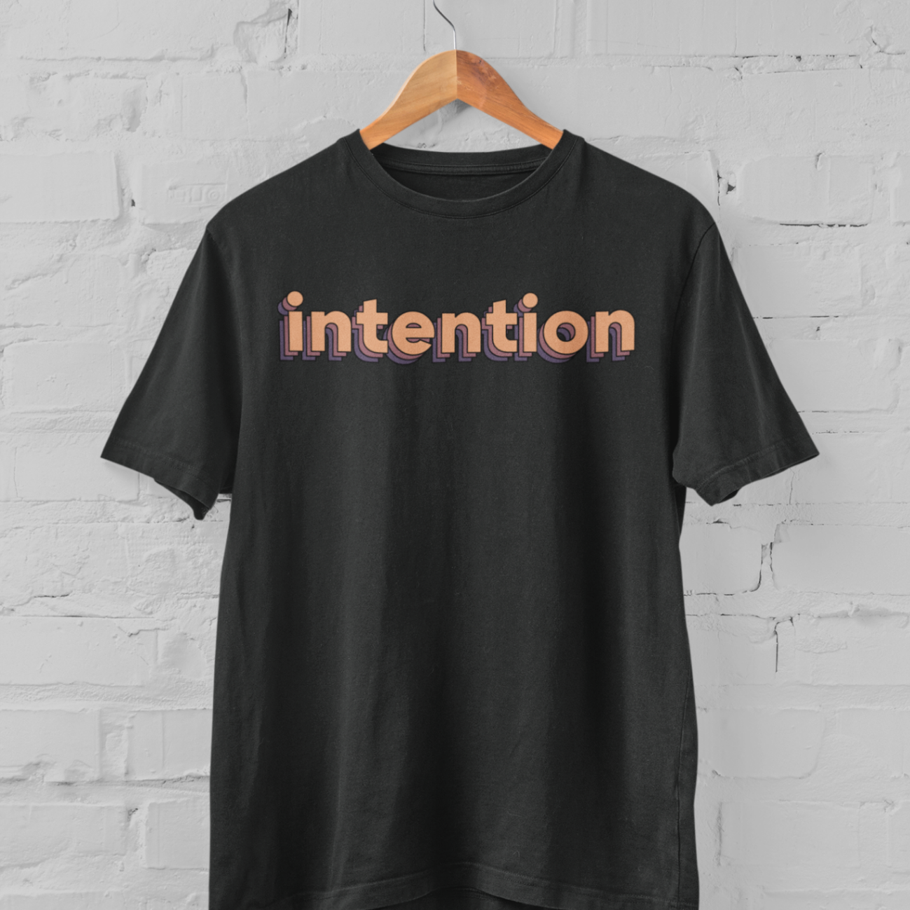 a vintage black t shirt with intention written in 3 D letters on a hanger in front of a white brick backround